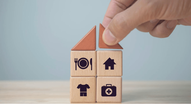 Hand places a roof piece on wooden blocks marked with home, dining, clothing, and medical icons.