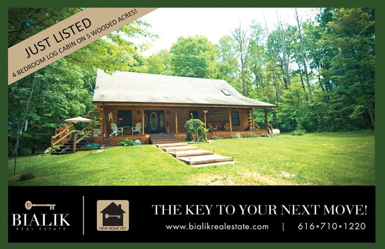 Log cabin with a broad porch in a wooded clearing, shown with a just-listed banner.