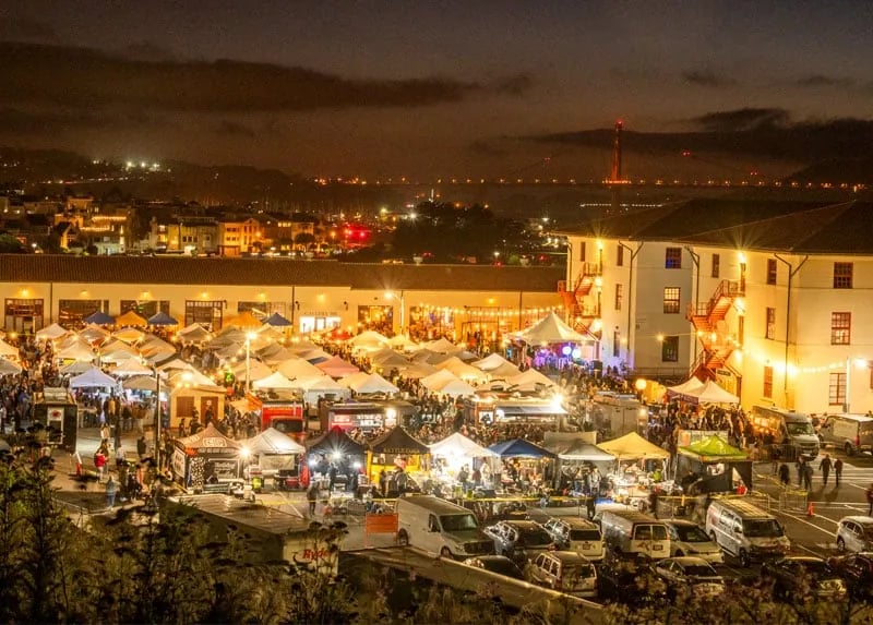 Aerial view of a bustling Fort Mason Night Market in San Francisco, showing dozens of white tents lit up, with the Golden Gate Bridge and city lights visible in the background.