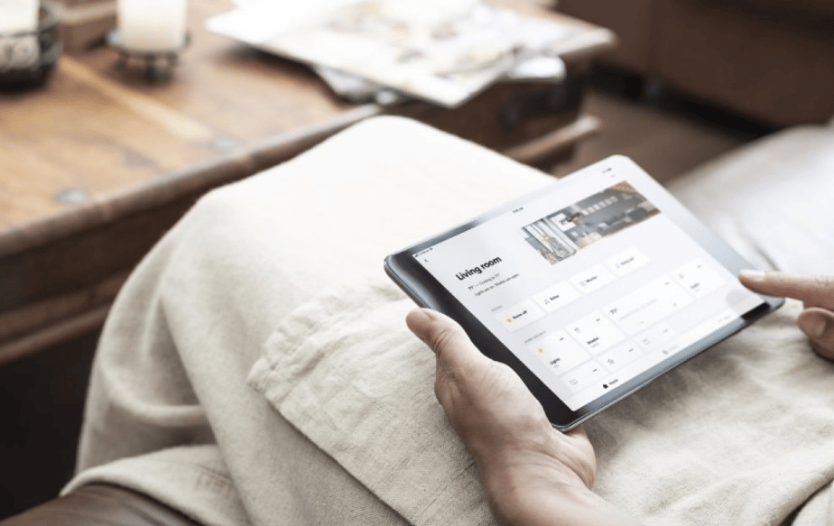 Person uses a tablet on a light sofa; the screen displays a living room interface.