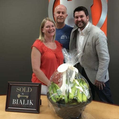 Three adults celebrate a sale beside a gift basket and framed SOLD sign.