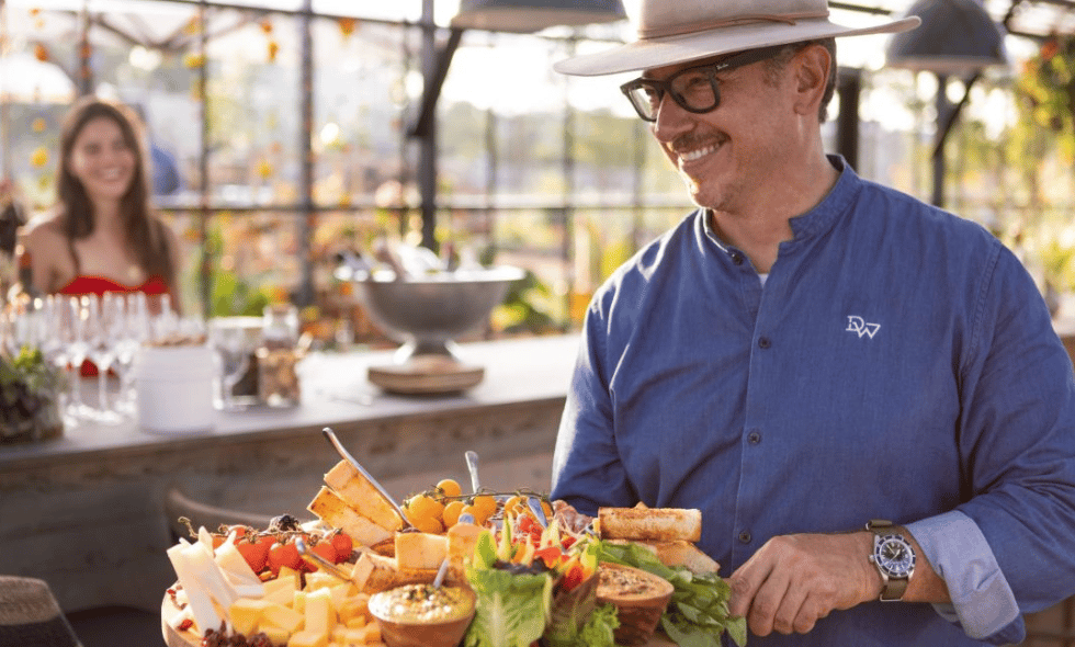 Man preparing a colorful cheese, fruit, and vegetable spread at a sunlit counter.