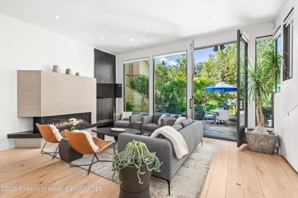 Modern living room with a gray sectional, linear fireplace, and glass doors opening to a leafy patio.
