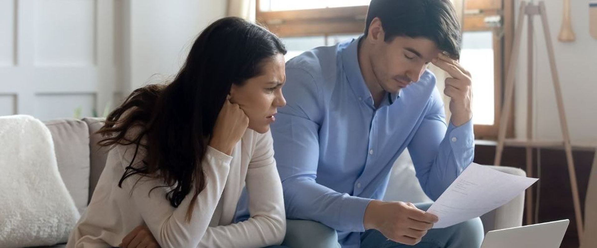 Worried couple reviews a document together on a living room sofa.