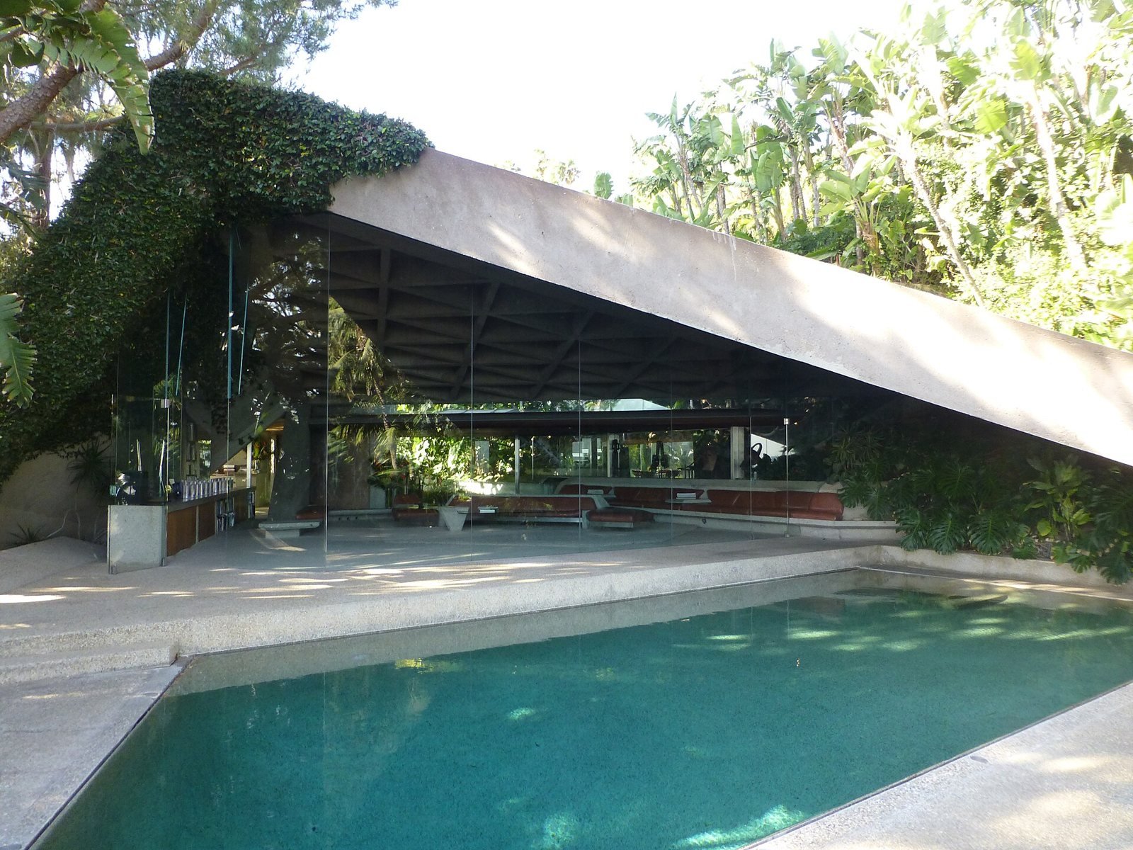 The Sheats-Goldstein Residence seen from across the pool, its angled concrete roof sloping down over a glass-walled living room, with planting growing over the roof edge.