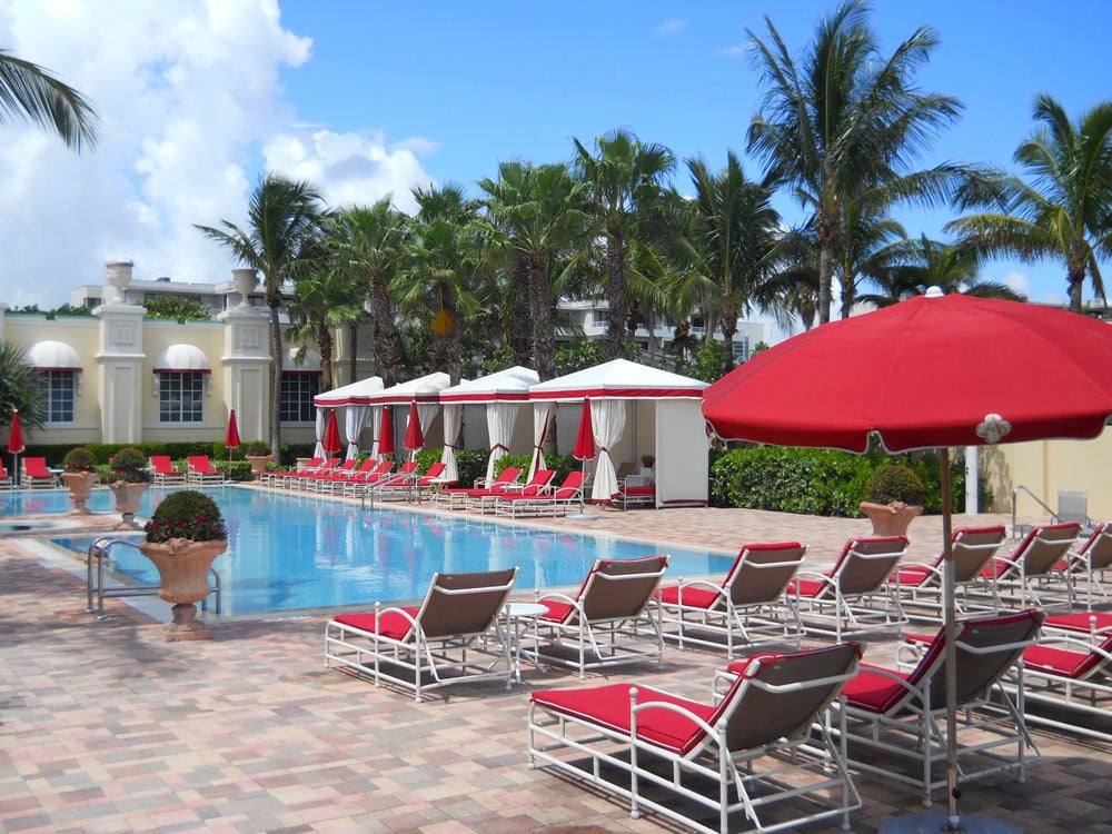 Resort pool with red-cushioned loungers, cabanas, palm trees, and a large red umbrella.
