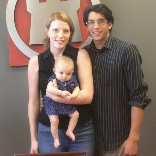 A couple stands with their baby in front of a large red-and-white sign.