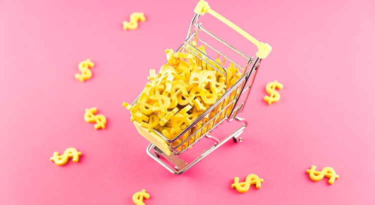 A small shopping cart filled with dollar signs on a pink background. This image likely represents financial concepts such as budgeting, expenses, or savings.  