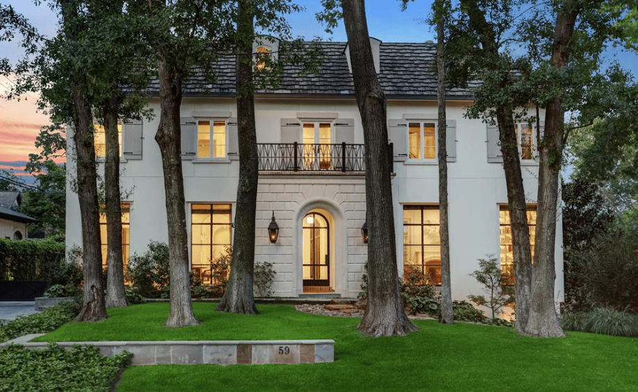 A three-story French-style mansion, with a white exterior, slate roof, balcony, and front lawn, nestled among trees.