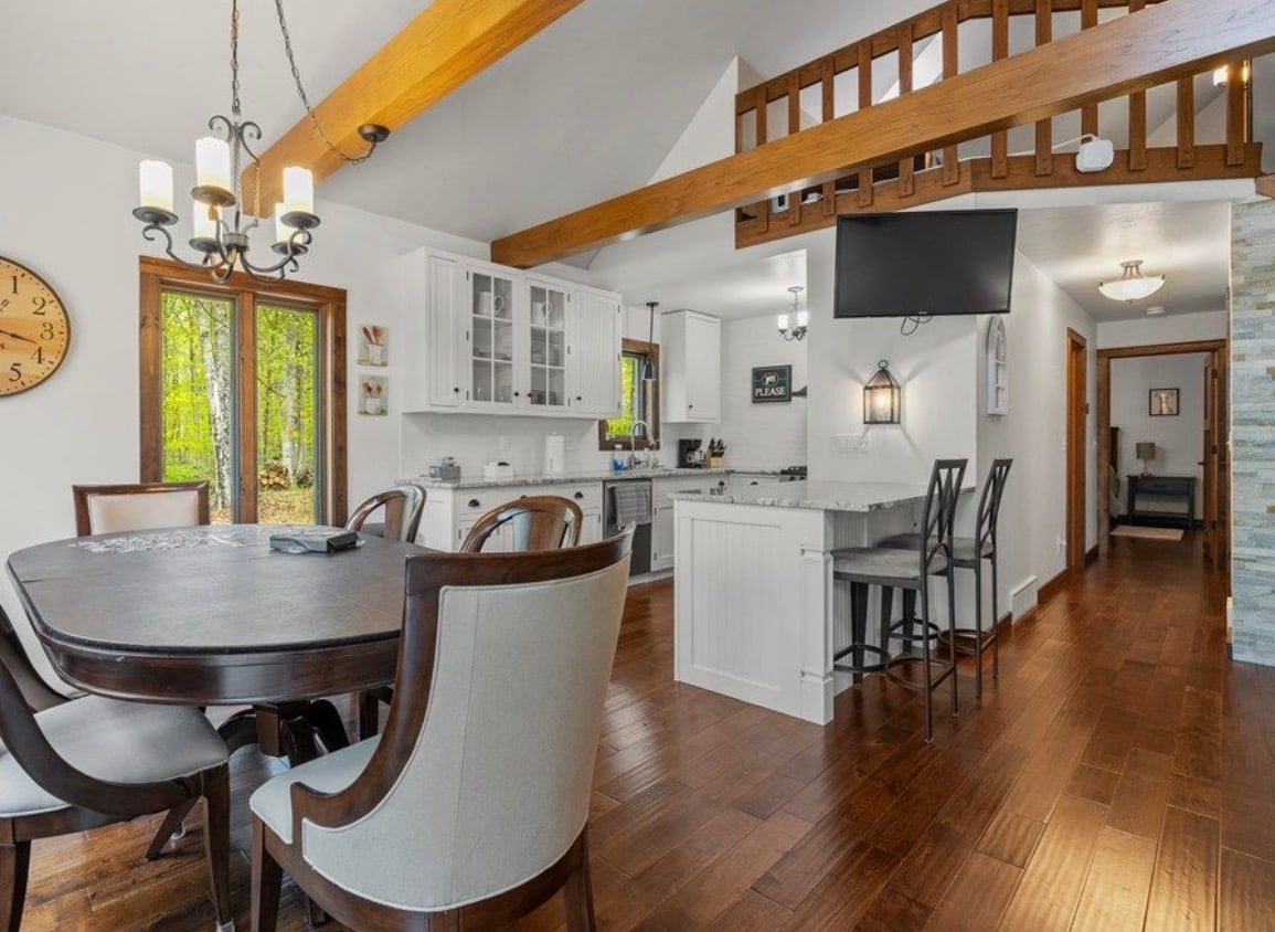 Open-plan kitchen and dining area with exposed wooden beams, a loft railing, and glass doors overlooking trees.