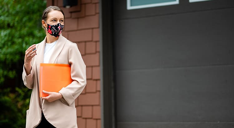 A person wearing a face mask holds a folder or notebook, standing outdoors near a building, possibly indicating a professional or academic context.