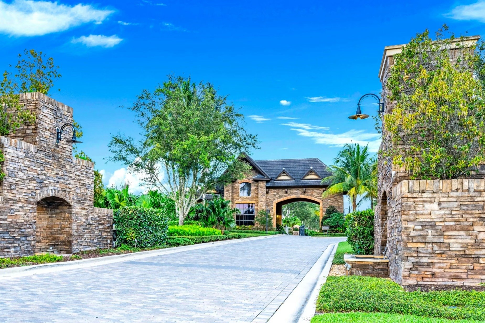 Gated community entrance with stone walls, an arched gatehouse, and tropical landscaping.