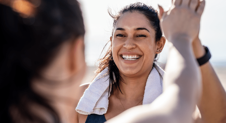 A woman with a towel around her neck smiles broadly while high-fiving another person. The image conveys a sense of joy and accomplishment outdoors.
