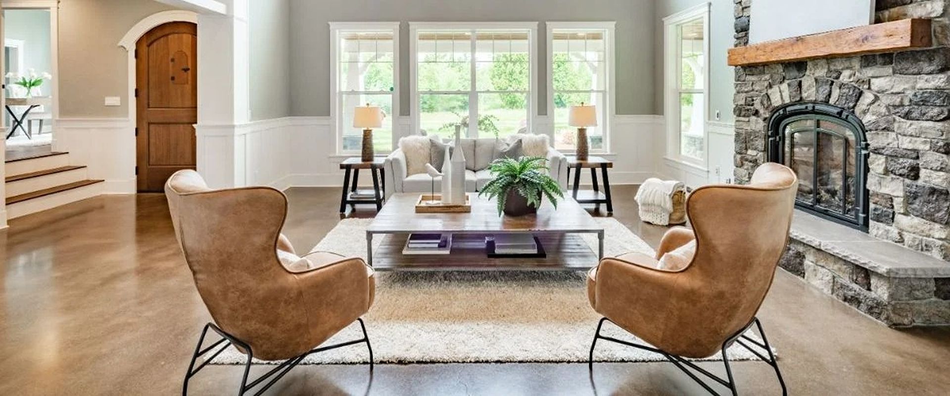 Staged living room with a stone fireplace, large windows, and tan armchairs facing a white sofa.