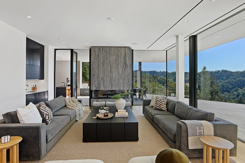 Staged living room with gray sofas, a stone fireplace, and wooded hills beyond floor-to-ceiling windows.