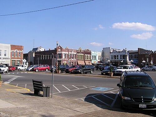 Downtown storefronts line a street with parked cars and a paved parking area.