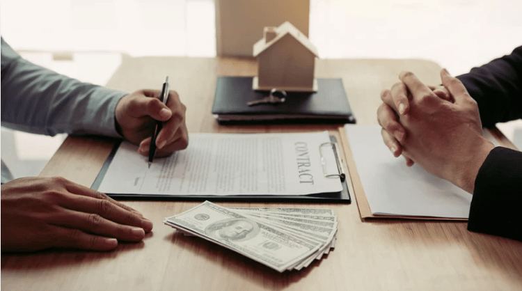 A person signs a real estate contract beside cash and a small model house.