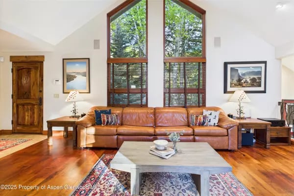 Living room with a leather sofa beneath tall peaked windows overlooking leafy trees.