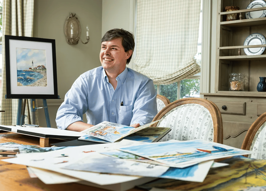 Artist seated at a table covered with colorful artwork in a bright studio.