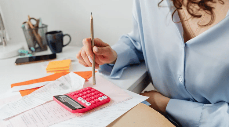 A person reviews paperwork with a pencil and pink calculator at a desk.