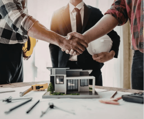 People shake hands over a miniature house as a real estate professional holds a folder.