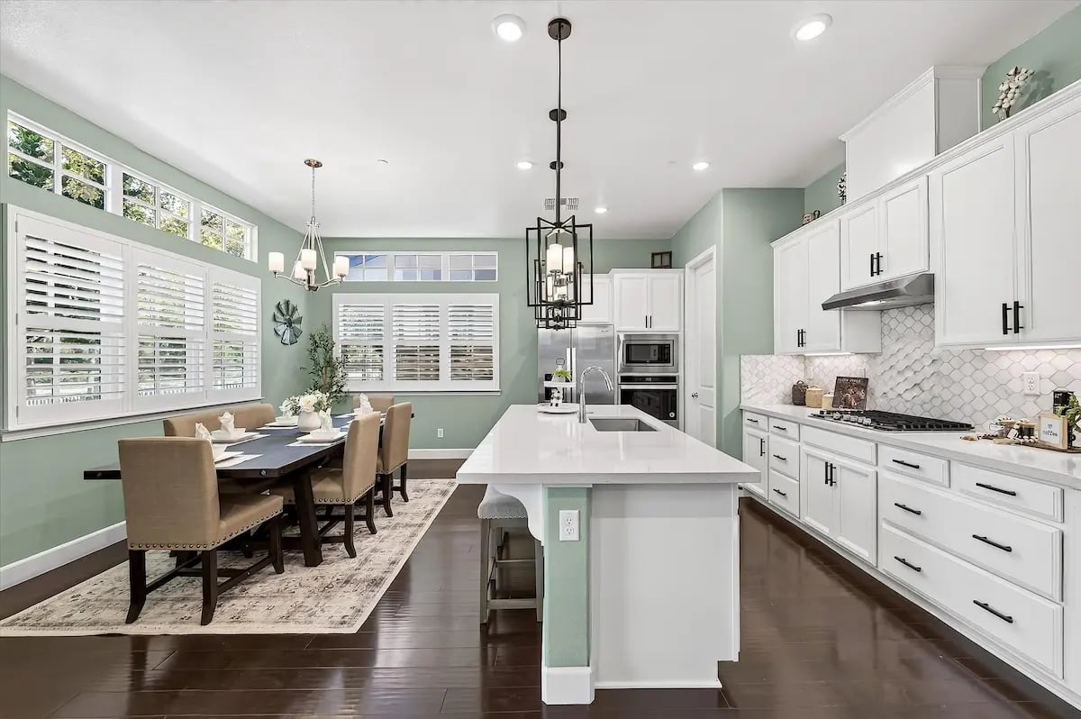 Bright kitchen and dining area with a white island, sage-green walls, and plantation shutters.