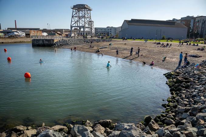 People wading in San Francisco Bay near rocky shoreline with historic crane structure