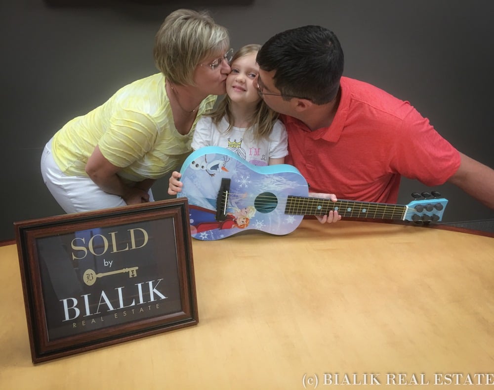 Two adults kiss a young girl holding a blue guitar beside a Bialik Real Estate sold sign.