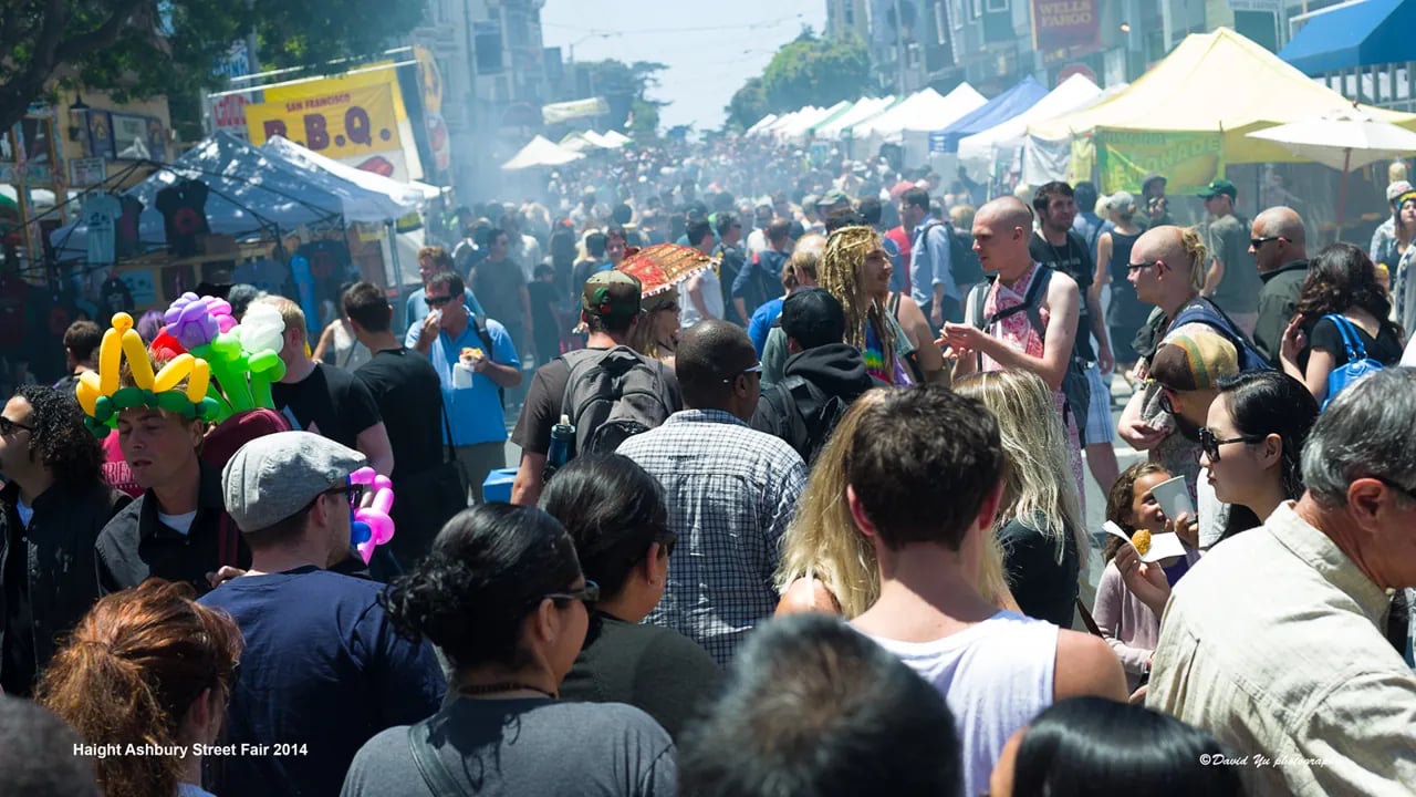 A large crowd of people walks past vendor booths and food tents at the Haight-Ashbury Street Fair.