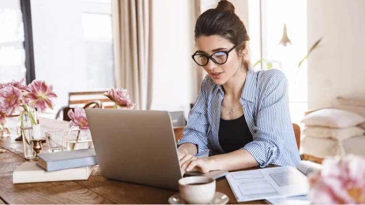Woman working on a laptop at a table with papers, books, and flowers.