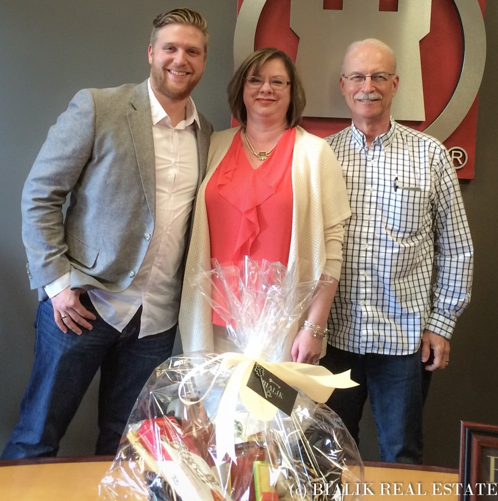 Three adults pose with a wrapped closing gift basket at a real estate office.