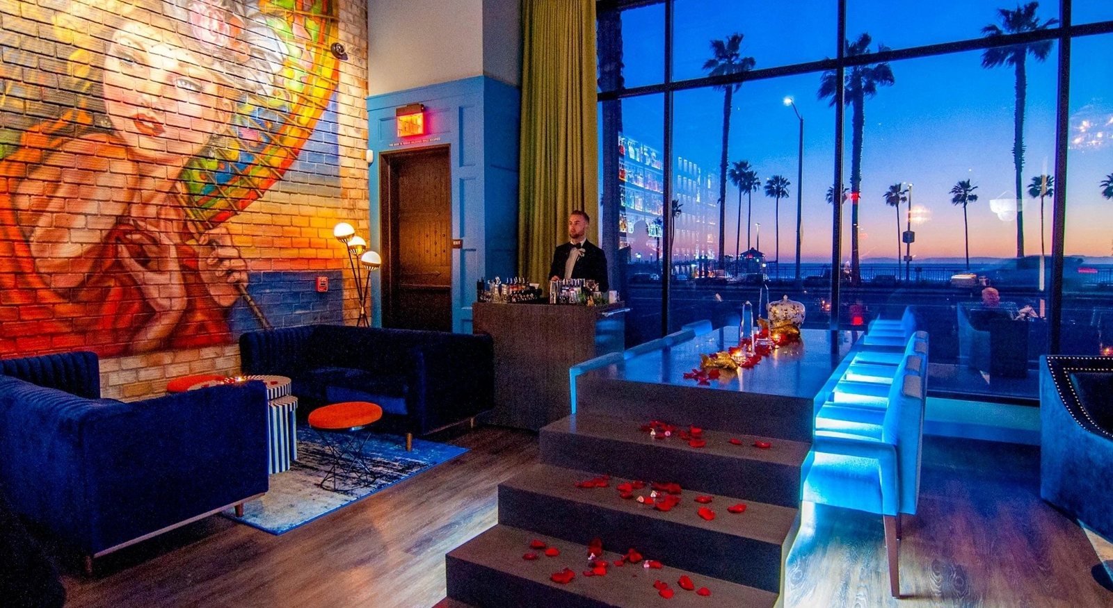 Graceland's lounge at dusk, with a painted brick mural, blue velvet seating and palm trees and ocean through the windows.