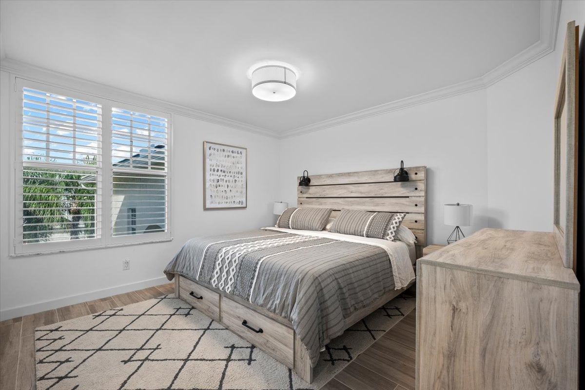 Bedroom with a gray wood bed, geometric rug, and shuttered windows overlooking greenery.