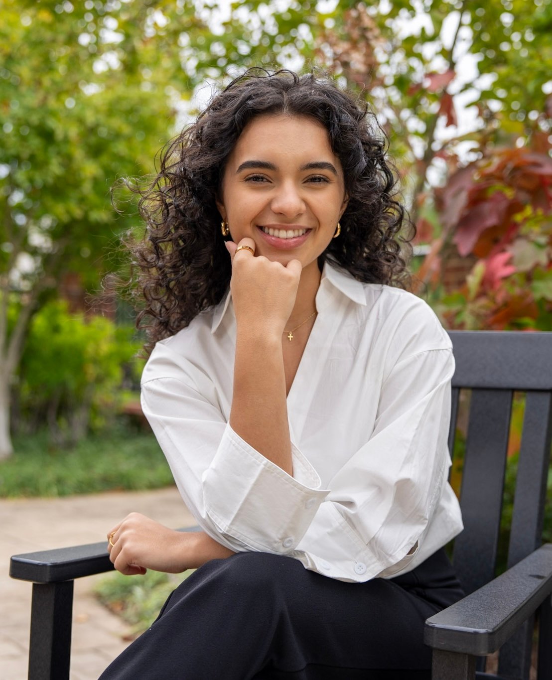 Michelle Isaacura smiling on a bench outdoors, wearing a white blouse and gold jewelry.