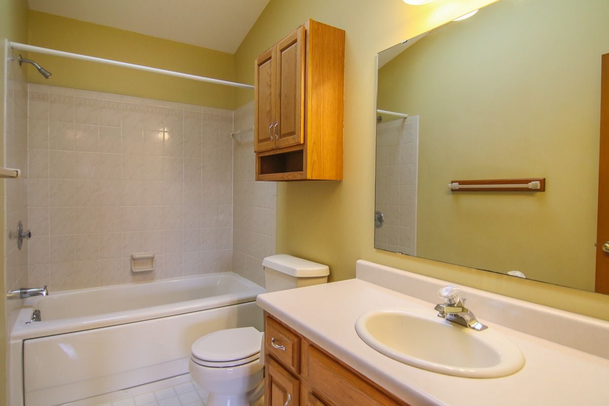 Bathroom with a white tub and tiled surround, toilet, and wood vanity with an oval sink.