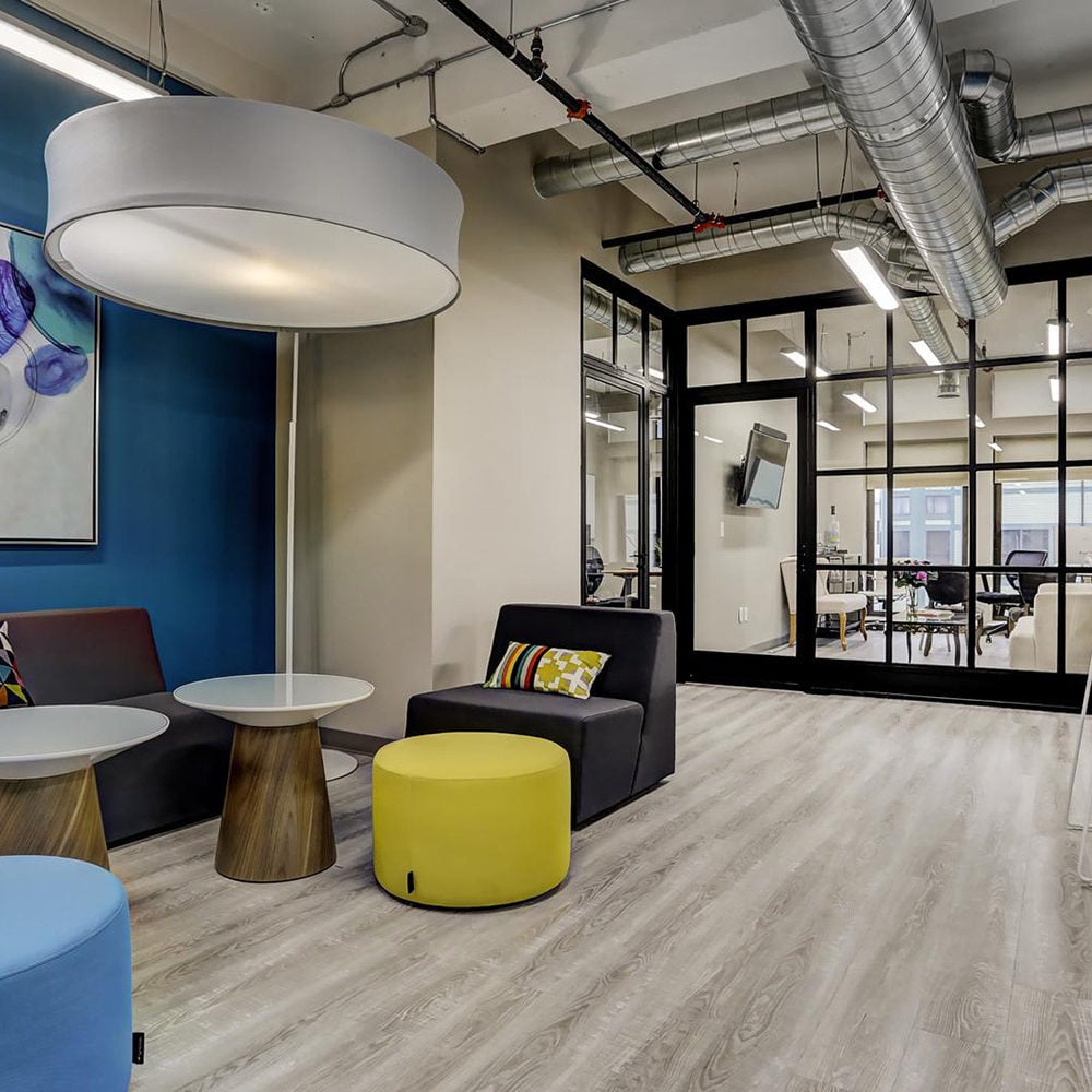 Office lounge with colorful seating and black-framed glass partitions opening onto a workspace.