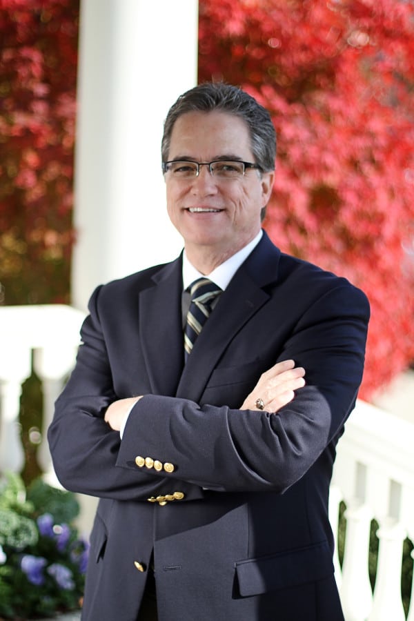 Man in a navy suit and striped tie standing with crossed arms against red autumn foliage.