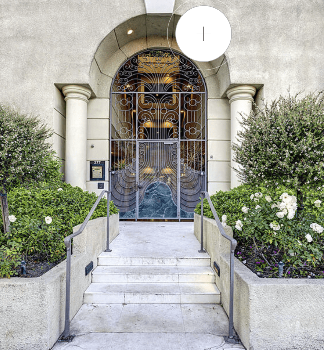 The entrance is marked by a wrought iron gate with a peacock design, surrounded by white flowers and green bushes.