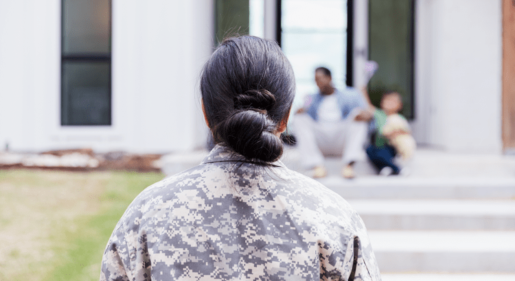 A person in military camouflage attire seen from behind, with their hair neatly tied in a bun. In the background, a family is visible, sitting on the steps of a house. 