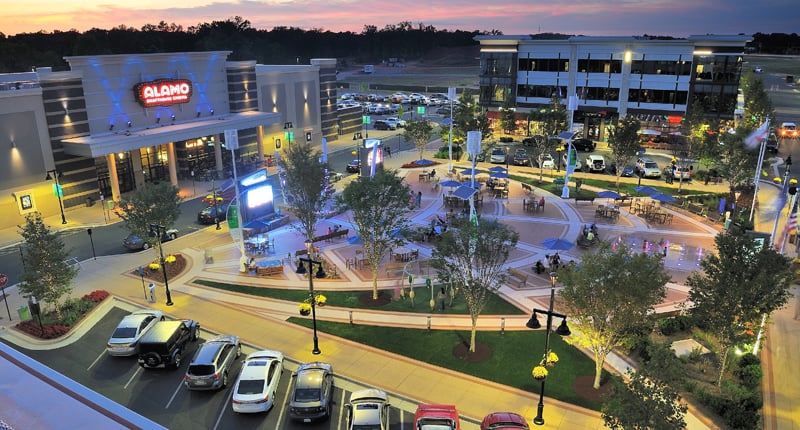 Evening view of the One Loudoun plaza in Ashburn, Virginia with outdoor dining umbrellas, string-lit walkways, the Alamo Drafthouse marquee and surrounding retail and office buildings