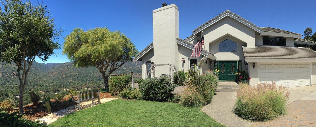 Two-story home with a green front door, landscaped lawn, and wooded hills behind it.