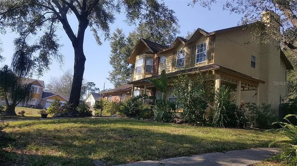 Two-story tan house with a covered porch, mature trees, and a landscaped front yard.