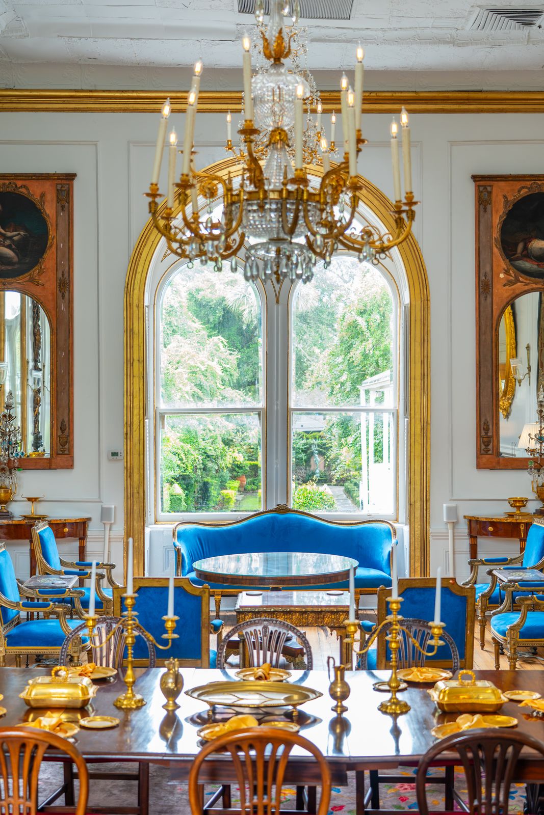 Formal dining room with a crystal chandelier, gilded arched window, and blue upholstered seating.