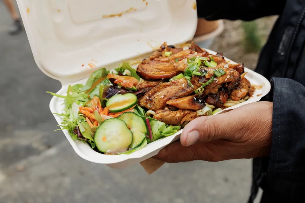 Person holding white takeout container with grilled meat over mixed green salad with cucumber slices, carrots, and garnishes