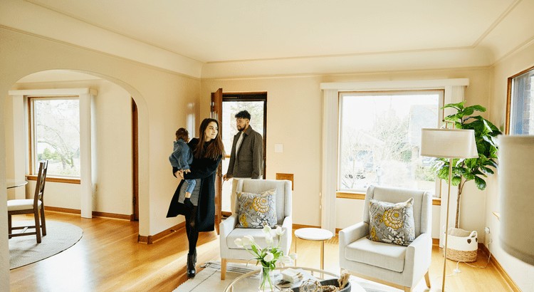 An interior scene with a couple being welcomed into a well-decorated living room by a real estate agent.