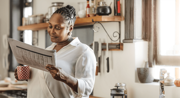 A woman in a bright kitchen reads a newspaper while holding a red coffee mug. She seems relaxed, surrounded by shelves with kitchen items.