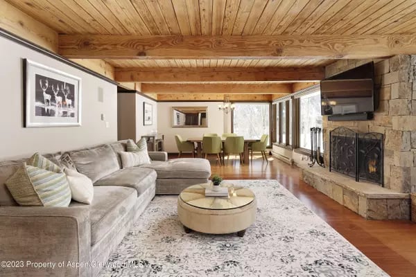Living room with a light sectional, exposed wood ceiling, stone fireplace, and dining area by large windows.