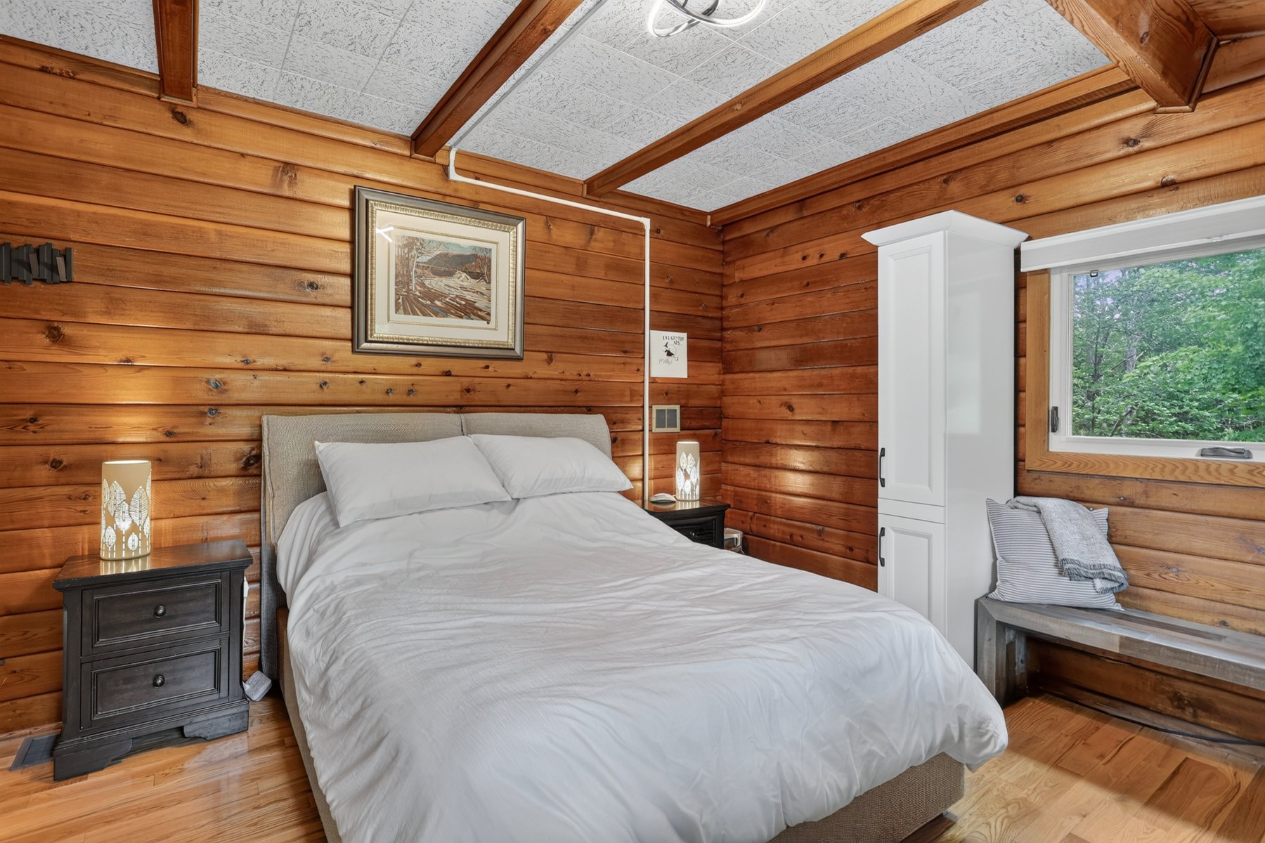 Bedroom with warm wood paneling, white bedding, and a window overlooking trees.