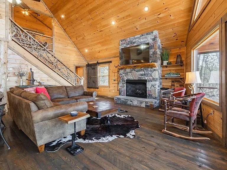 Wood-paneled living room with a vaulted ceiling, stone fireplace, staircase, and sectional sofa.