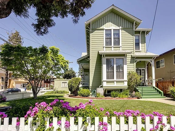 Light green two-story house with white trim, a front porch, and pink flowers behind a white picket fence.